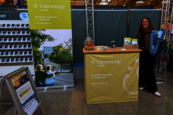 Das Bild zeigt Katrin Reinhardt, Leitung und Koordinatorin des AHPB Lebenswege in Ludwigshafen am Infostand im Globus in Oggersheim, Sie steht zwischen dem Fischstand und Scan&go an der rechten Seite der Theke. Dahinter das RollUp der Lebenswege. Der Stand wirbt für ehrenamtliche Hospizbegleitung 
