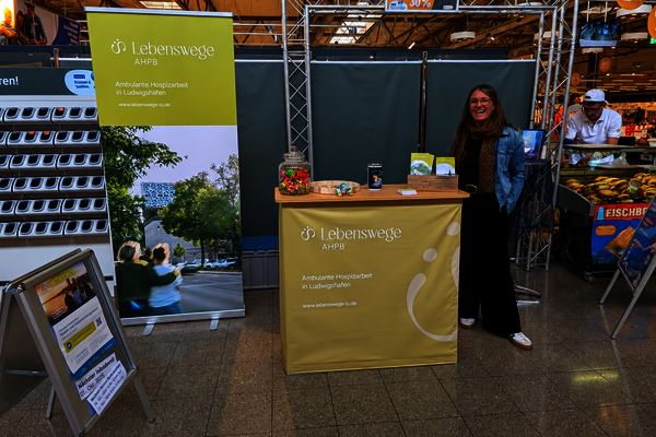 Das Bild zeigt Katrin Reinhardt, Leitung und Koordinatorin des AHPB Lebenswege in Ludwigshafen am Infostand im Globus in Oggersheim, Sie steht zwischen dem Fischstand und Scan&go an der rechten Seite der Theke. Dahinter das RollUp der Lebenswege. Der Stand wirbt für ehrenamtliche Hospizbegleitung 